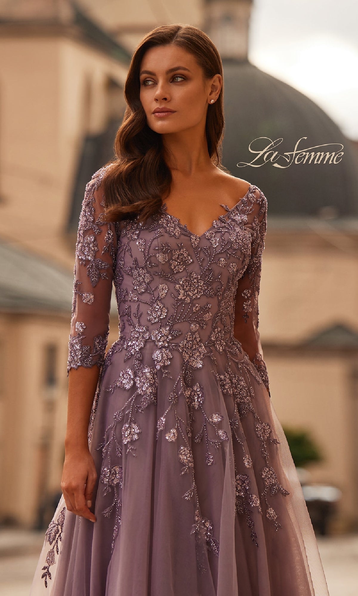 A woman in the La Femme 32538 A-Line Tulle Formal Dress with three-quarter sleeves and beaded lace appliques stands outdoors before blurred classical architecture.